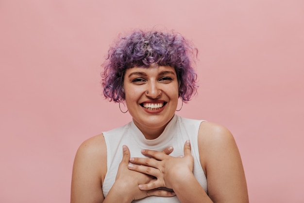 Insolito giovane donna con i capelli viola in bianco superiore sorridente. Bella signora con acconciatura luminosa in posa sul rosa.