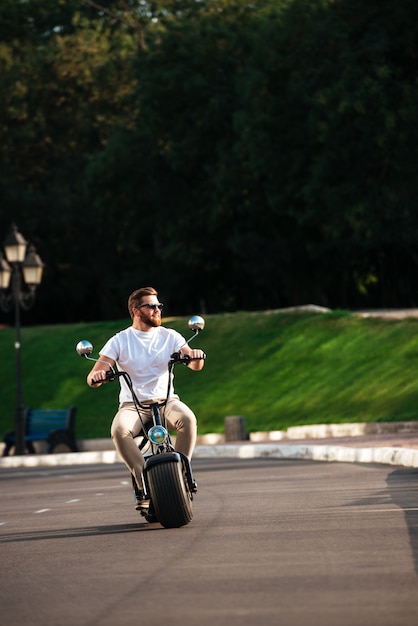 Integrale dell'uomo barbuto sorridente in occhiali da sole cavalca sulla moto moderna all'aperto e guardando lontano