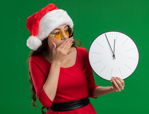 interessato giovane bella ragazza che indossa santa cappello e occhiali tenendo e guardando l'orologio tenendo la mano sulla bocca isolato su sfondo verde