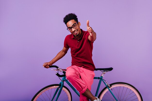 Interessato modello maschio africano in posa. Elegante ragazzo nero con gli occhiali seduto sulla bicicletta blu.