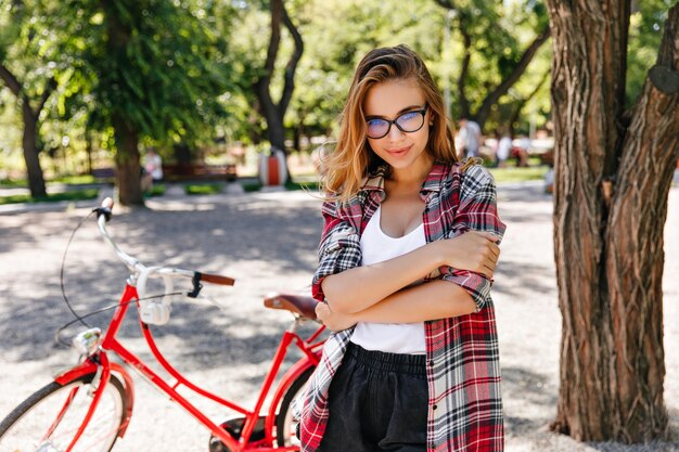 Interessato ragazza caucasica in camicia a scacchi in posa nel parco in una giornata estiva. Donna bionda di buon umore che si rilassa dopo un giro in bicicletta.