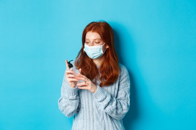 Inverno, covid-19 e concetto di distanza sociale. Giovane donna dai capelli rossi con maschera facciale pulisce le mani con antisettico, disinfetta con disinfettante per le mani, in piedi su sfondo blu