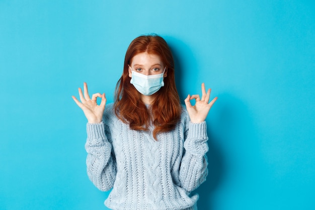 Inverno, covid-19 e concetto di distanza sociale. Soddisfatta giovane donna rossa in maschera facciale che mostra bene, gesti ok e sembra soddisfatta, in piedi sfondo blu.