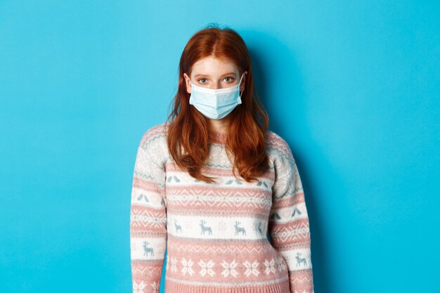 Inverno, covid-19 e concetto di quarantena. Giovane ragazza rossa in maglione e maschera per il viso che fissa la telecamera, in piedi su sfondo blu.