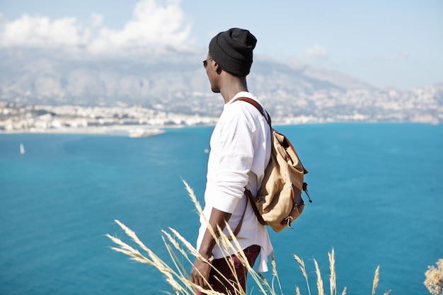 Irriconoscibile giovane turista afroamericano alla moda che gode del bel tempo estivo e del meraviglioso paesaggio marino intorno a lui, mentre in piedi sulla cima della montagna durante un'escursione nell'area tropicale del resort