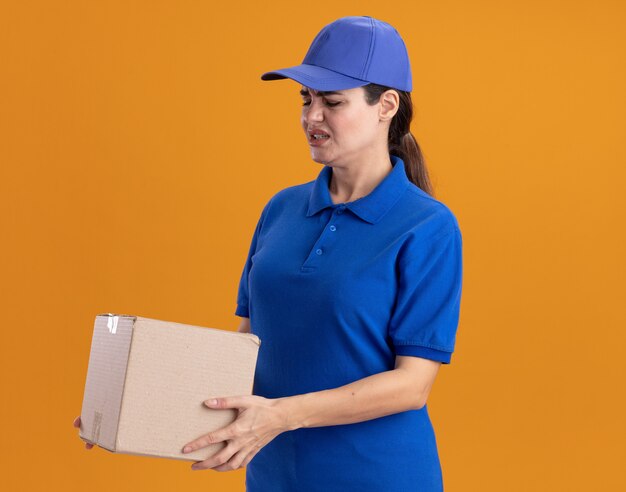 Irritata giovane donna delle consegne in uniforme e berretto in piedi in vista di profilo tenendo e guardando cardbox isolato sulla parete arancione con spazio di copia