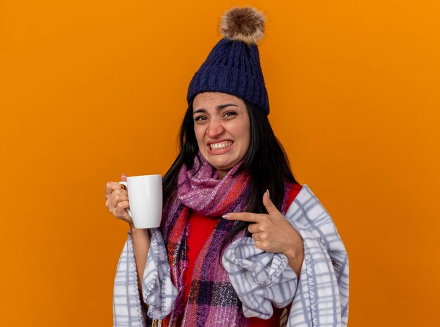 Irritata giovane ragazza malata caucasica che indossa cappello invernale e sciarpa avvolta in plaid che tiene e che indica alla tazza di tè isolata sulla parete arancione con lo spazio della copia