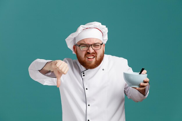 Irritato giovane chef maschio che indossa occhiali uniforme e cappuccio che mostra una ciotola vuota con un cucchiaio in esso guardando la fotocamera che mostra il pollice in giù isolato su sfondo blu