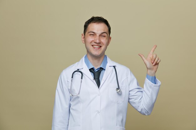 Irritato giovane medico maschio che indossa accappatoio medico e stetoscopio intorno al collo guardando la fotocamera facendo gesto di pistola rivolto verso l'alto isolato su sfondo verde oliva