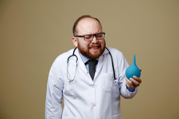 Irritato giovane medico maschio che indossa occhiali camice da laboratorio e stetoscopio intorno al collo tenendo clistere con gli occhi chiusi isolati su sfondo verde oliva