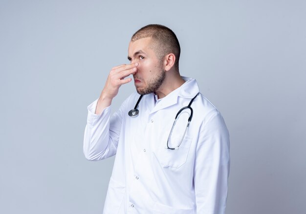 Irritato giovane medico maschio indossa abito medico e stetoscopio intorno al collo tenendo il naso guardando il lato isolato su bianco