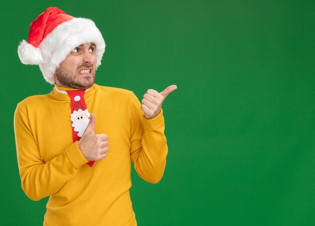 Irritato giovane uomo caucasico che indossa cappello di Natale e cravatta guardando e indicando il lato che mostra il pollice in alto isolato su sfondo verde