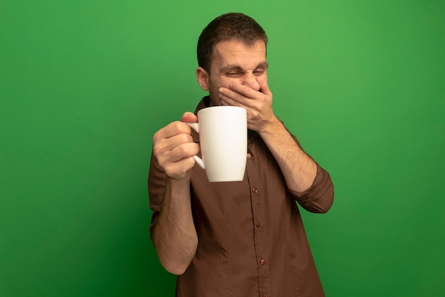 Irritato giovane uomo caucasico isolato su azienda verde e guardando la tazza di tè mantenendo la mano sulla bocca sfondo con copia spazio