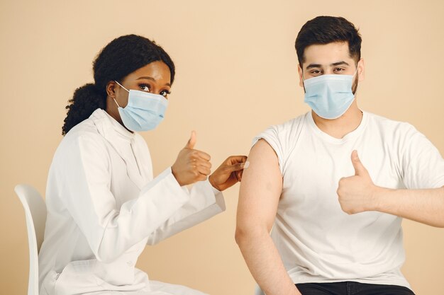 Isolato, sfondo beige. Operaio medico donna africana e uomo in maschere che mostrano i pollici in su. Vaccinazione contro il covid.