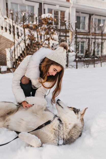 Ispirata signora bianca con cappello che scherza con husky sulla neve. Foto all'aperto di ridere giovane donna che gioca con il suo cane in cortile.