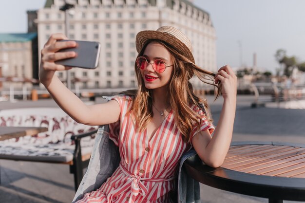 Ispirata signora europea con cappello di paglia che gioca con i suoi capelli e fa selfie. Foto all'aperto dell'adorabile ragazza bianca in abito a righe che cattura maschera di se stessa sulla città.