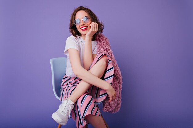 Ispirato donna caucasica in maglietta bianca e scarpe sognante in posa sulla sedia Sorridente ragazza affascinante con capelli mossi seduta su sfondo viola