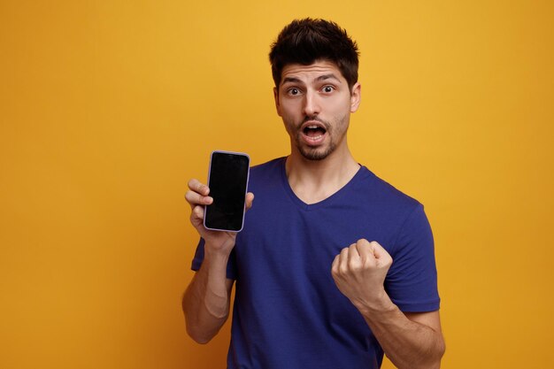 Ispirato giovane bell'uomo guardando la fotocamera che mostra il telefono cellulare tenendo il pugno in aria su sfondo giallo