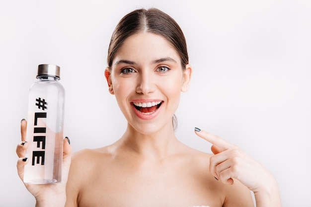 Istantanea del modello sorridente attraente sul muro bianco. La ragazza senza trucco indica la bottiglia, dimostrando che l'acqua è vita.