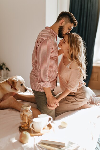 Istantanea di mattina di fine settimana di coppia a letto con colazione e cane sdraiato nelle vicinanze. Il giovane barbuto inala il profumo dei capelli delle sue amate ragazze.