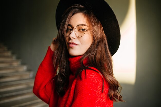 Istantanea di una giovane donna attraente con un cappello a tesa larga sul muro delle scale. La ragazza in occhiali e maglione rosso sta sorridendo.