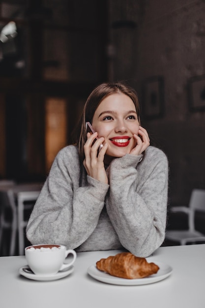 Istantanea di una ragazza affascinante che sorride sognante e parla al telefono Una donna con un maglione grigio ha ordinato un croissant e una tazza di caffè nella caffetteria