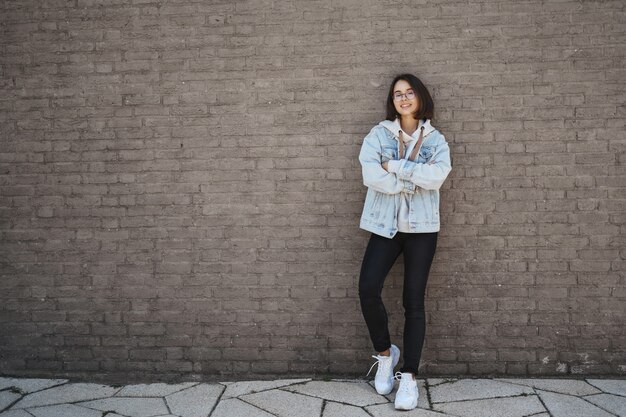 Istruzione, stile di vita e concetto di generazione moderna. Fiduciosa giovane donna felice in giacca di jeans, occhiali, muro di mattoni magro e sorridente, mani incrociate sul petto fiduciosa, inviando CV trovato lavoro.