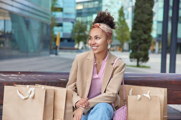 L'acquirente positivo della donna dai capelli ricci si riposa dopo aver effettuato acquisti nel centro commerciale vestito con abiti alla moda circondato da borse della spesa guarda con gioia le pose su uno sfondo urbano sfocato