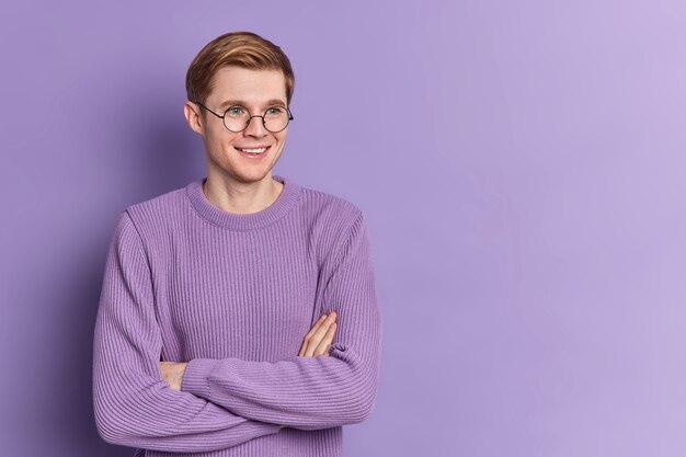 L'adolescente bello allegro dell'uomo tiene le braccia conserte guarda pensieroso da parte ha un sorriso gentile indossa pose di base del ponticello