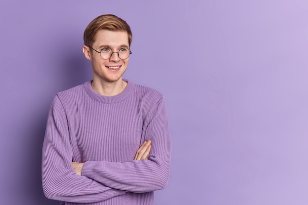 L'adolescente bello allegro dell'uomo tiene le braccia conserte guarda pensieroso da parte ha un sorriso gentile indossa pose di base del ponticello