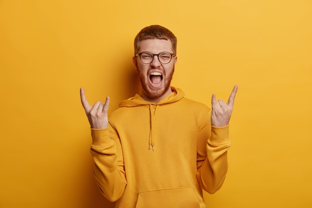 L'adolescente emotivo eccita di gioia, fa il gesto del rock n roll, porta vibrazioni positive, gode di un concerto fantastico, ascolta la musica preferita, vestito con una felpa, isolato su un muro giallo. Linguaggio del corpo