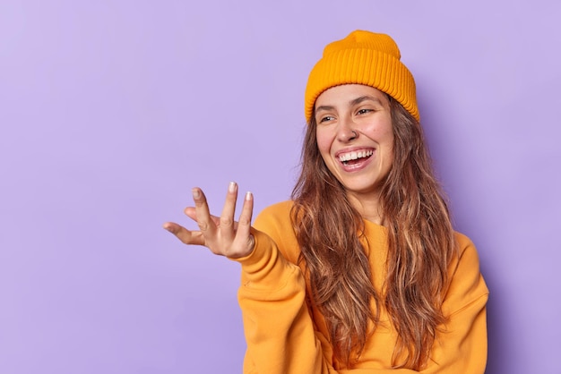 L'adolescente felice alza la mano guarda con gioia qualcosa di divertente indossa cappello e maglione posa su sfondo viola con uno spazio vuoto per i tuoi contenuti pubblicitari. Concetto di emozioni umane positive