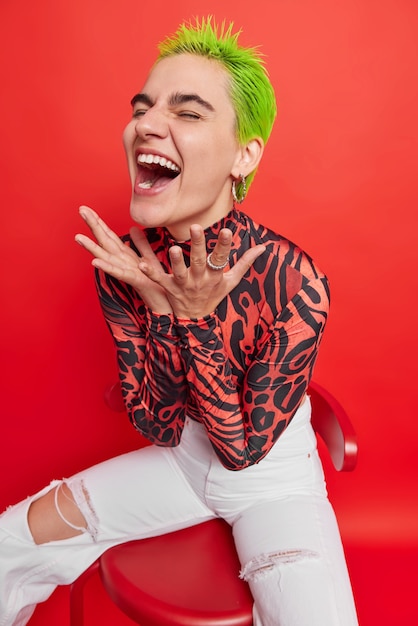 L'adolescente felice esclama dalla gioia si siede comodamente sulla sedia reagisce emotivamente su una cosa divertente ha un aspetto non standard capelli tinti verdi vestiti con abiti casual isolati su un muro rosso vivo