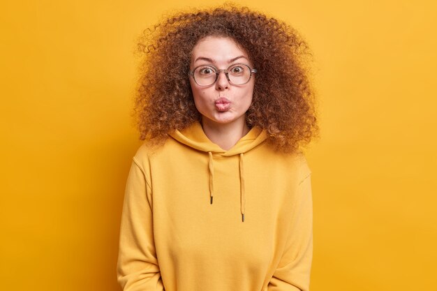 L'adolescente romantica mantiene le labbra arrotondate vuole baciare il fidanzato mentre ha un appuntamento indossa grandi occhiali trasparenti e felpa casual isolata sul muro giallo. Momento di tenera relazione