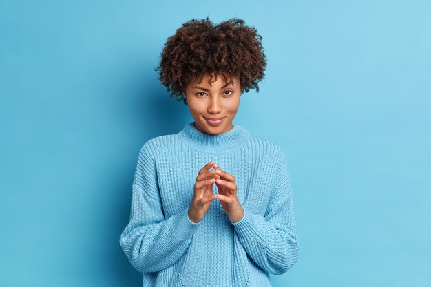 L'adorabile modella dalla pelle scura guarda misteriosamente alla telecamera ha un'espressione ingannevole mentre intriga qualcosa e le dita campeggianti fanno piani stand all'interno dello studio indossa un maglione blu casual