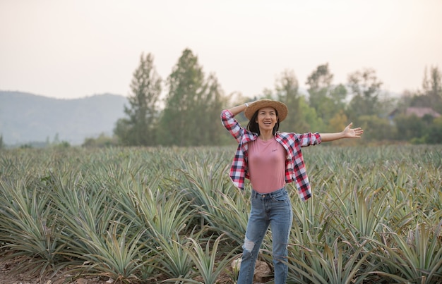 L'agricoltore femminile asiatico vede la crescita dell'ananas in fattoria, giovane donna graziosa del coltivatore che sta sul terreno coltivabile con le braccia alzate su felicità euforica gioiosa.