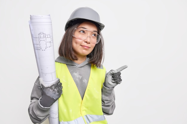 L'architetto femminile asiatico serio indossa indumenti protettivi di sicurezza del casco e occhiali trasparenti tiene il progetto indica via sullo spazio della copia mostra la direzione al cantiere. Concetto di settore