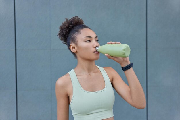 L'atleta femminile con un'espressione motivata ha una pausa di allenamento beve acqua fredda dalla bottiglia vestita in pose ritagliate contro il muro grigio