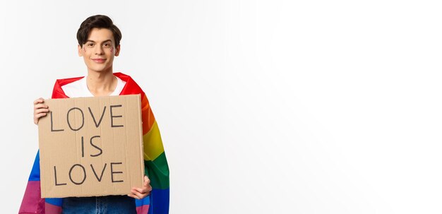 L'attivista gay sorridente che tiene il segno L'amore è amore per la parata dell'orgoglio lgbt che indossa la bandiera arcobaleno su sfondo bianco