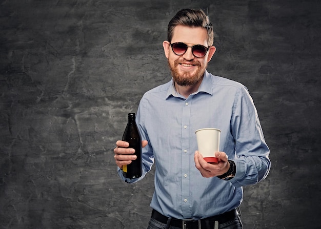 L'elegante maschio hipster barbuto tiene una tazza di caffè di carta e una birra artigianale in bottiglia.