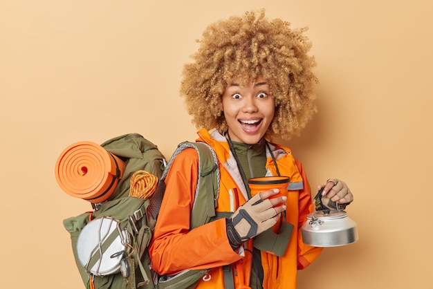 L'escursionista femminile positivo posa con gli utensili ride felicemente vestito con una giacca arancione porta lo zaino conduce uno stile di vita attivo essendo di buon umore isolato su sfondo beige ha una vacanza in montagna
