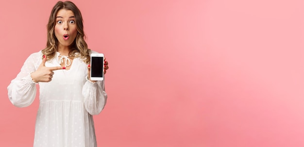 L'hai vista un'app fantastica Una bella ragazza bionda impressionata ed eccitata in abito bianco consiglia di scaricare l'applicazione visita il negozio online indicando il telefono cellulare e fissa la fotocamera elettrizzata