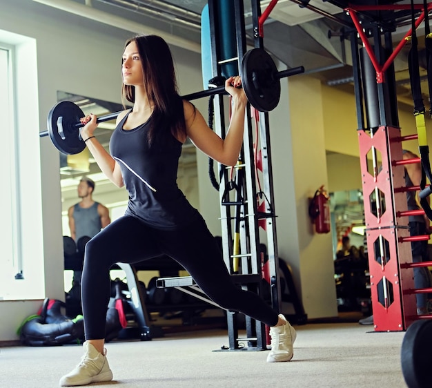L'immagine completa del corpo di una donna bruna sportiva che fa squat con bilanciere in una palestra.