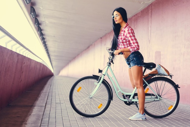 L'immagine del corpo intero della donna sportiva in pantaloncini di jeans e maglietta in pile che posa su una bicicletta in un tunnel con pareti rosa.