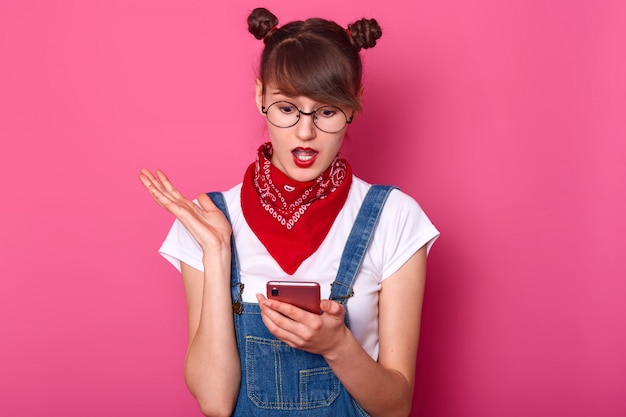 L'immagine dell'adolescente scioccato tiene il cellulare moderno, ha un messaggio con cattive notizie dalla sua amica, perplessa. La ragazza turbata guarda lo smartphone, vede che il suo ragazzo non è online. Concetto di tecnologia.