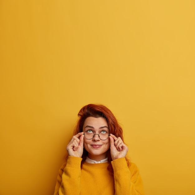 L'immagine dell'interno della donna caucasica rossa premurosa tiene le mani sulla montatura degli occhiali, cerca di vedere qualcosa sopra, ha un'espressione curiosa e felice