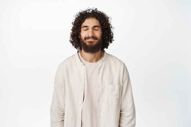 L'immagine dell'uomo ispanico calmo e rilassato chiude gli occhi sorridendo, respirando e riposando in piedi in abiti casual su sfondo bianco