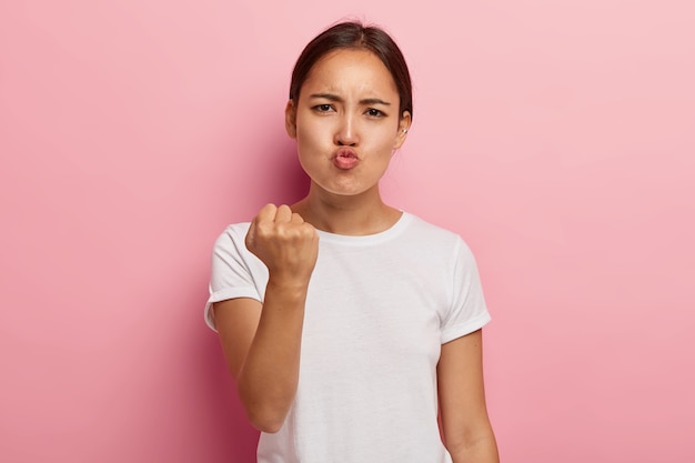 L'immagine della donna asiatica insoddisfatta arrabbiata stringe il pugno con dispiacere, tiene le labbra piegate, fa l'espressione del viso oltraggiato, indossa abiti bianchi, ti minaccia, isolato sul muro rosa.