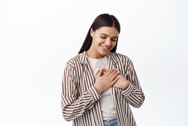 L'immagine della donna carina e tenera che si tiene per mano sul cuore che guarda in basso e che sorride sincera si sente grata e soddisfatta in piedi su uno sfondo bianco