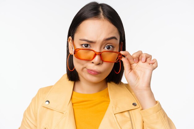 L'immagine della giovane donna asiatica alla moda che guarda da sotto gli occhiali da sole con la faccia scettica di incredulità esprime il dubbio in piedi su sfondo bianco